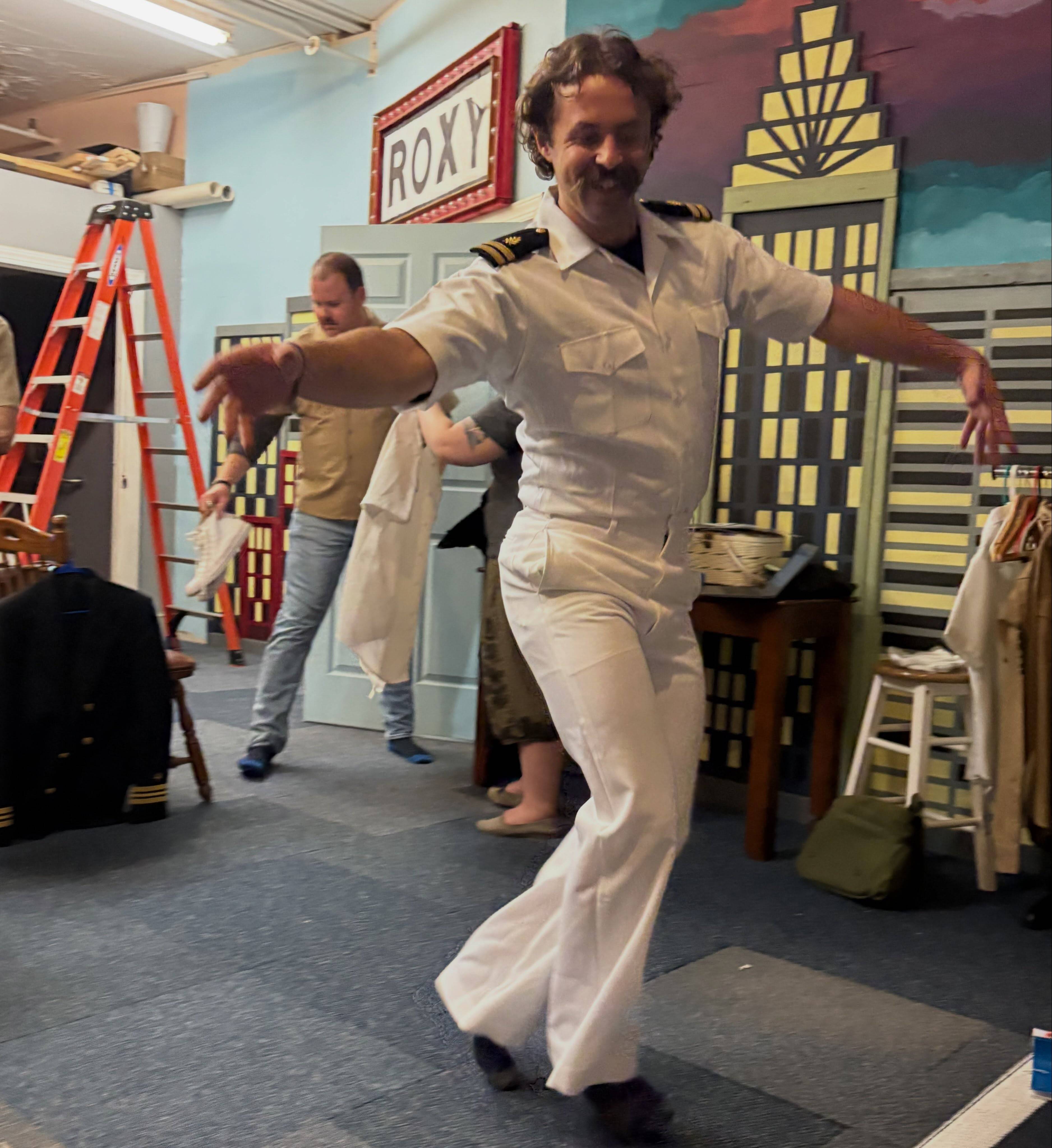 The same actor in the white Navy uniform strikes a dance pose mid-spin in the rehearsal room.