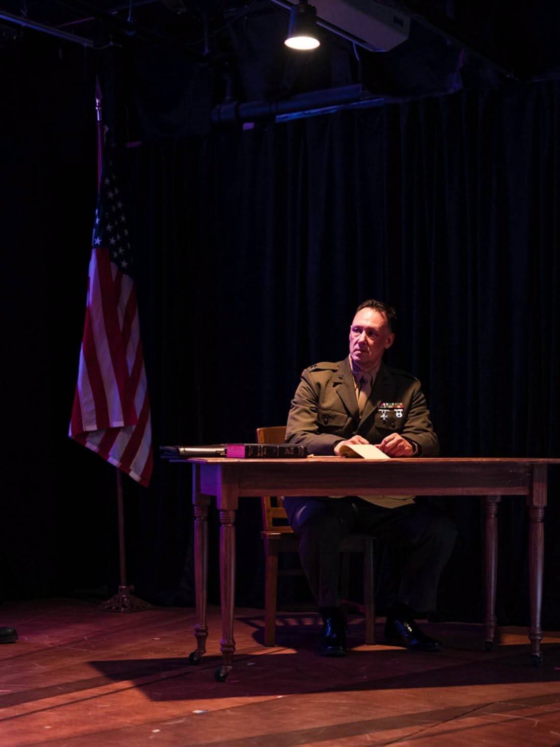 A uniformed official sits alone beneath a spotlight and United States flag.