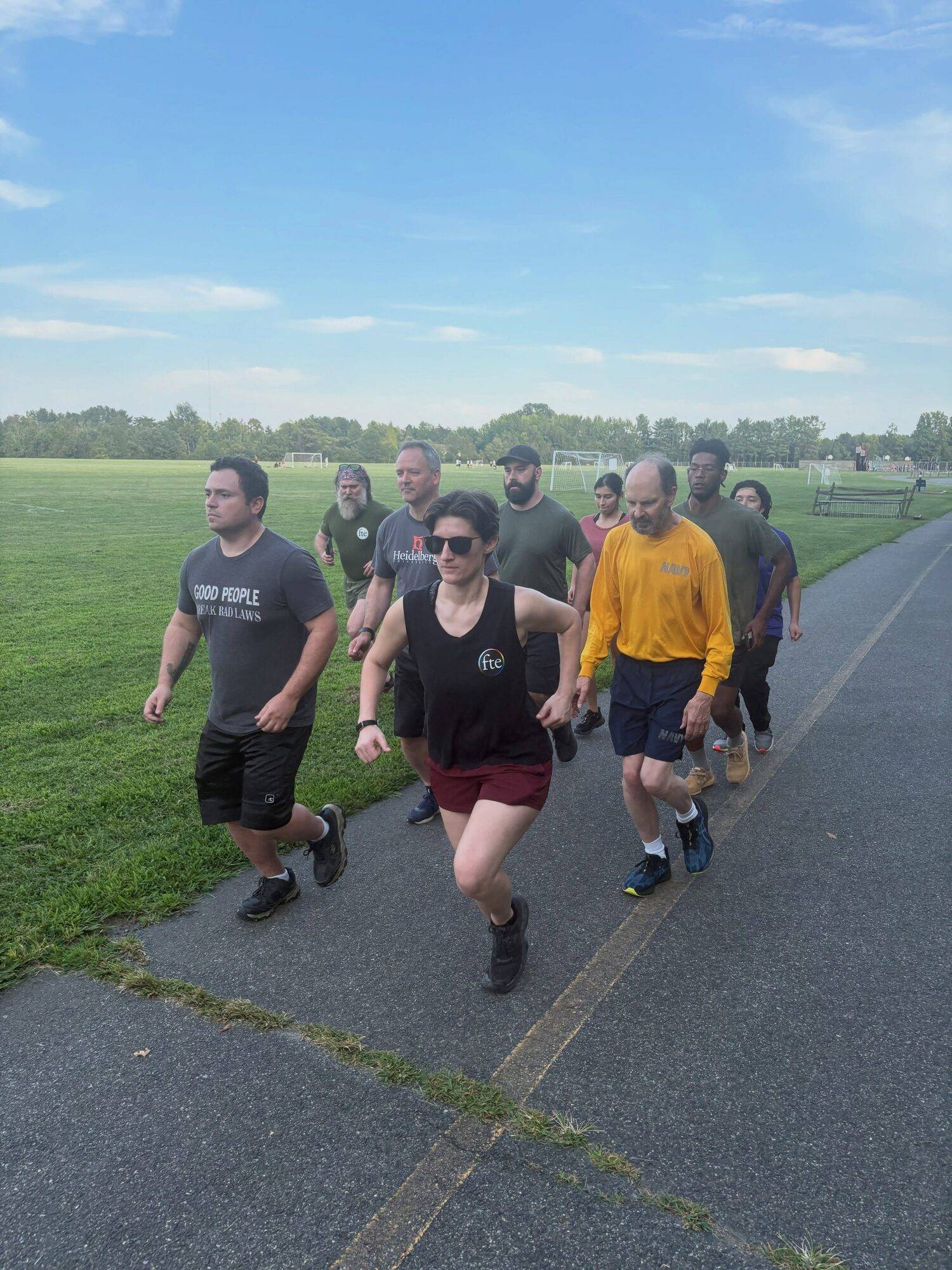 The group runs in formation along a paved track past open playing fields, the runner in front in a Fredericksburg Theatre Ensemble tank top, one behind in a T-shirt reading “Good people break bad laws” and another in a yellow Navy shirt.
