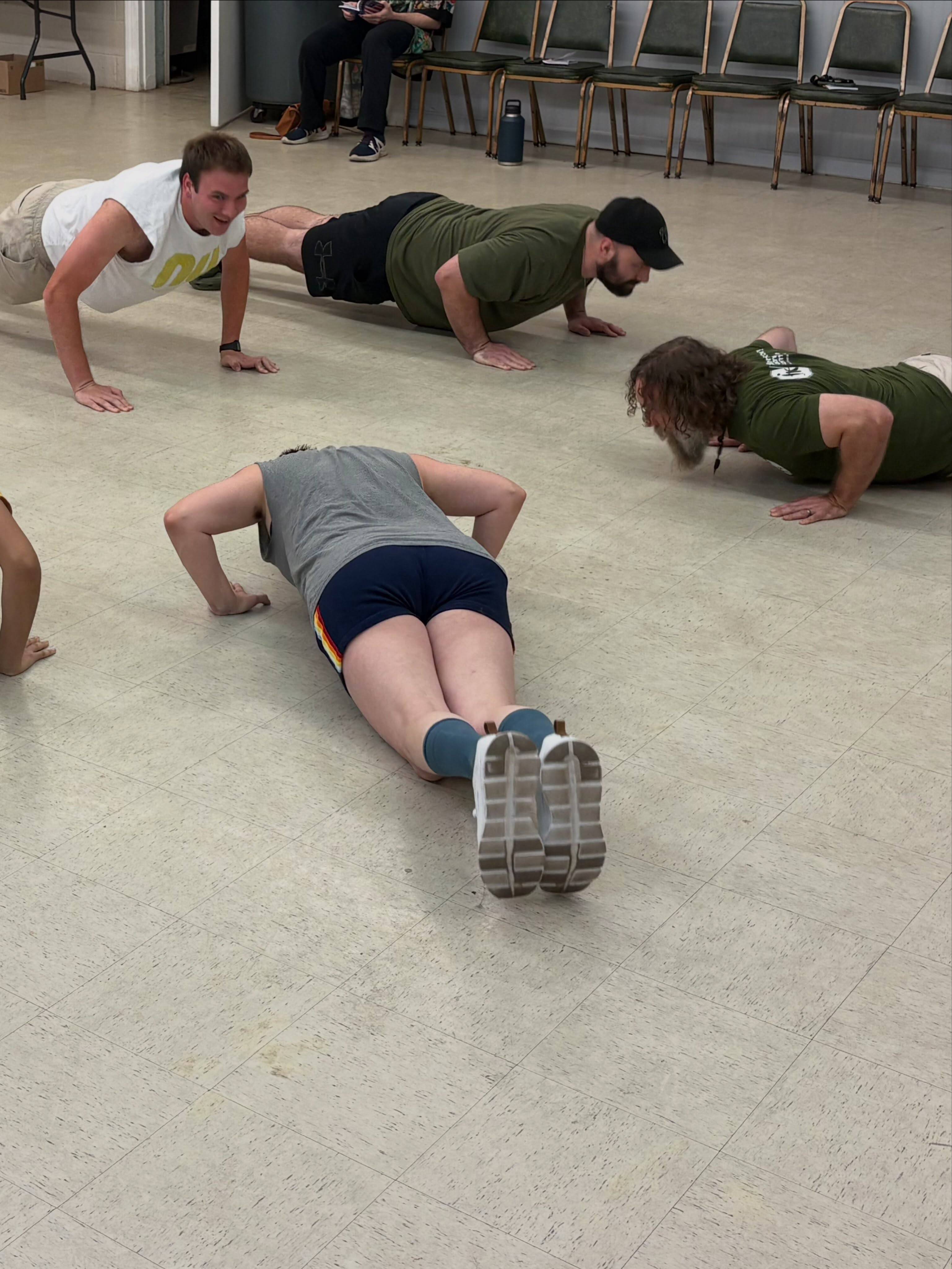 Four cast members hold a push-up position on the rehearsal room floor, one grinning at the camera.