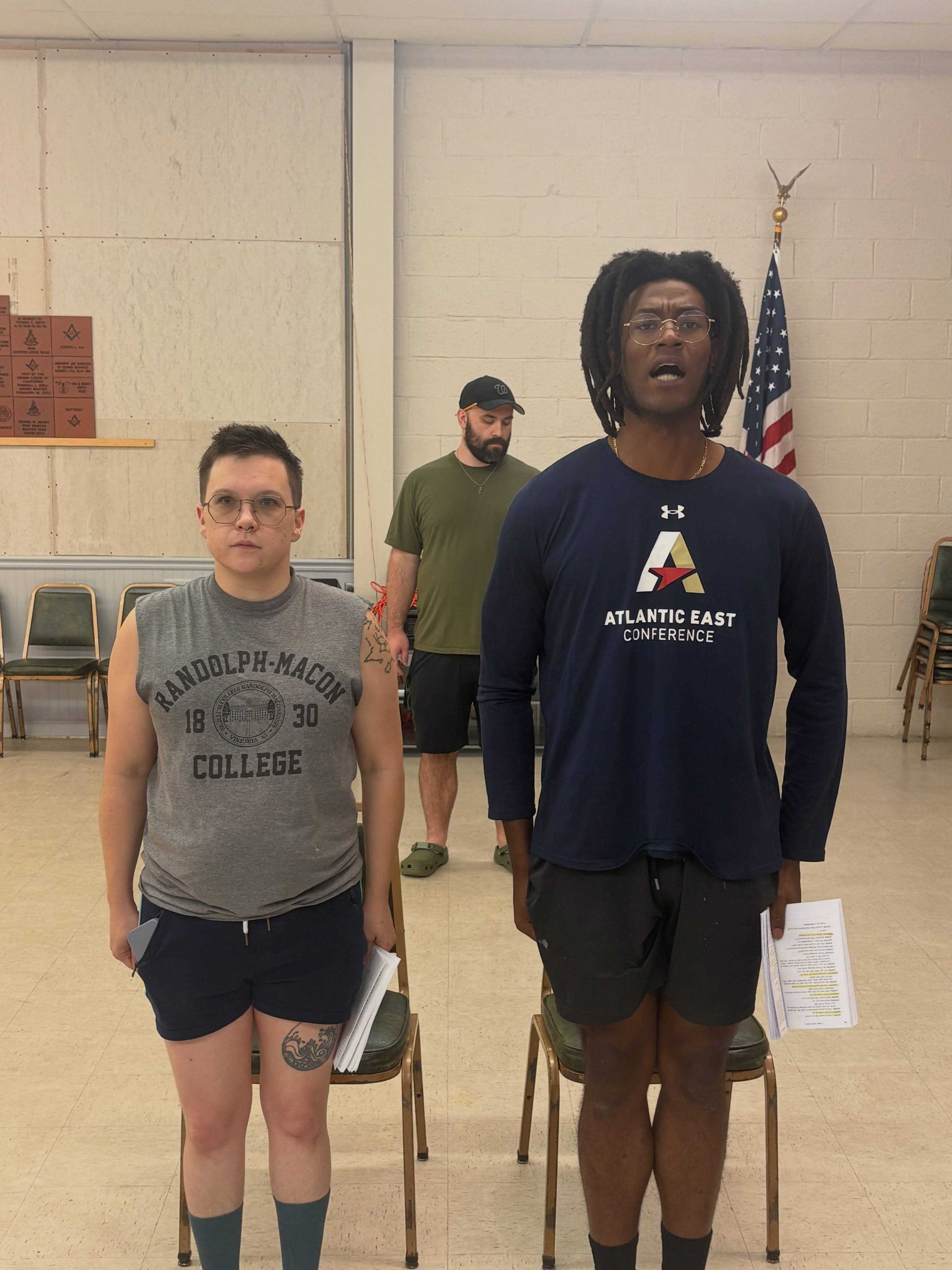 Two cast members stand at attention shouting, an American flag on the wall behind them.