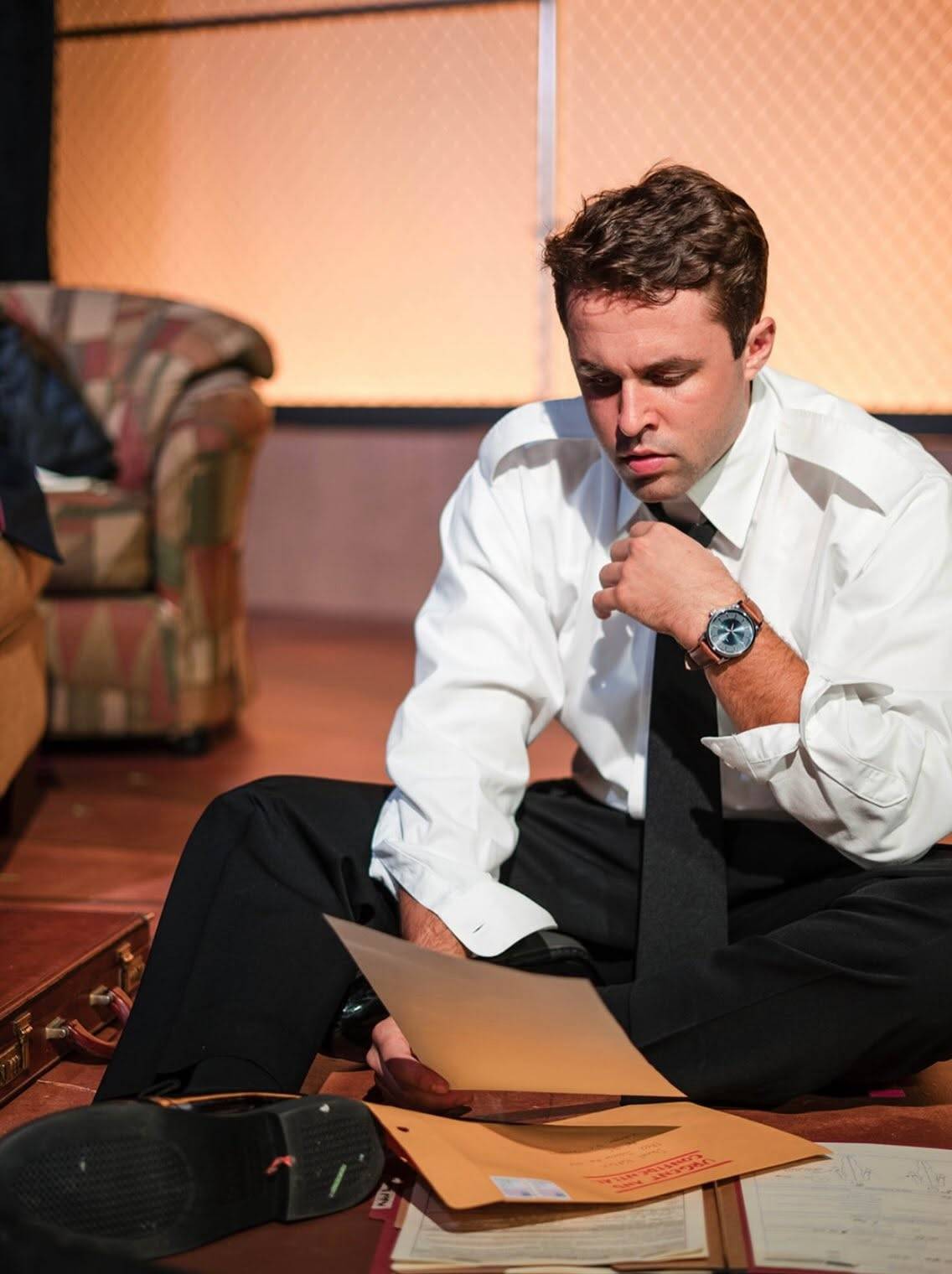 A performer in a white dress shirt with the sleeves rolled up, a loosened black tie and black trousers sits on the stage floor reading a sheet of paper, with an open leather briefcase, manila envelopes and scattered files around them. A patterned armchair and a chain-link wall lit warm orange stand behind.