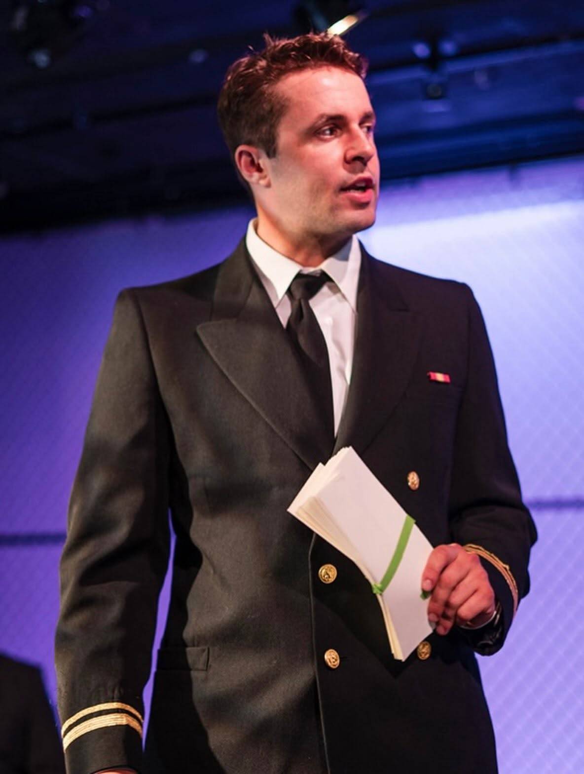 A performer in a dark double-breasted naval service dress coat with gold buttons and gold sleeve braid stands speaking, holding a bound stack of papers tied with a green ribbon. The chain-link wall behind them is lit deep purple.