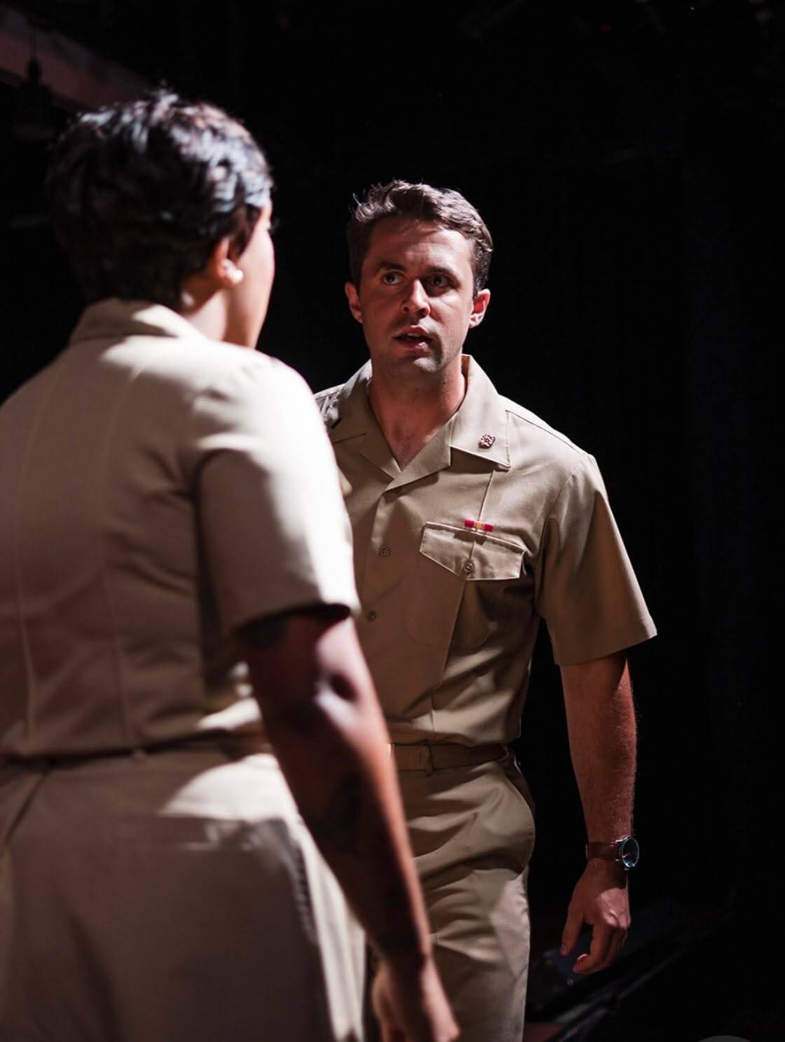 Two performers in tan service uniforms face each other in a pool of light against a black stage. One stands in the foreground with their back to us, out of focus; the other looks at them mid-sentence, a ribbon bar above the shirt pocket and a wristwatch on the arm hanging at their side.