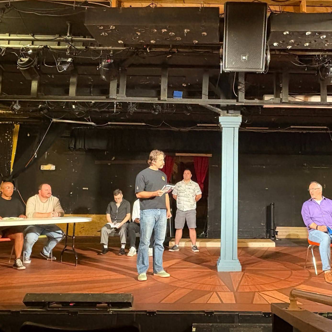A rehearsal on a wood-inlay stage under exposed lighting rig and a hanging speaker: one performer stands centre reading from a script, another stands behind, and several more sit watching from a folding table and chairs on either side.