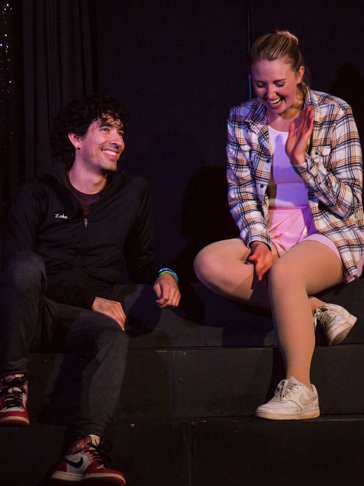 Two performers sit side by side on the stage steps, laughing together.
