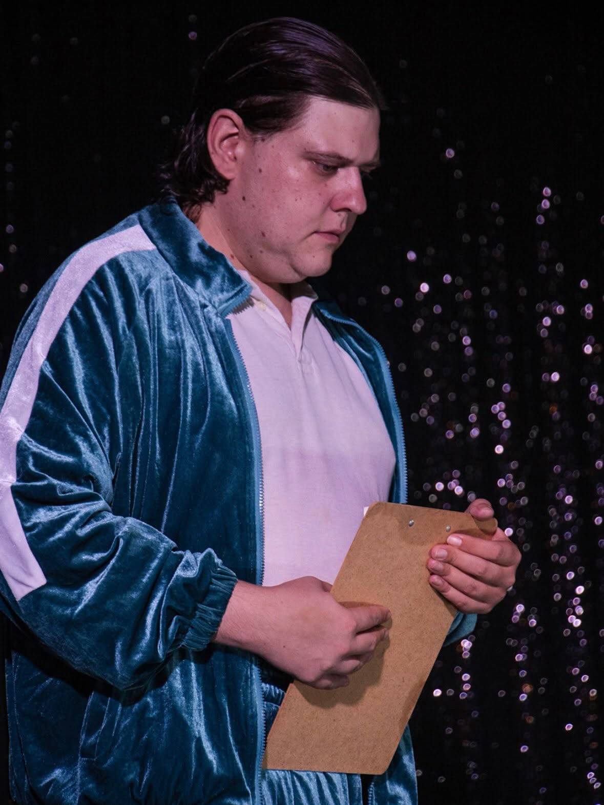 A performer in a teal velvet tracksuit studies a clipboard in front of a sparkling backdrop.