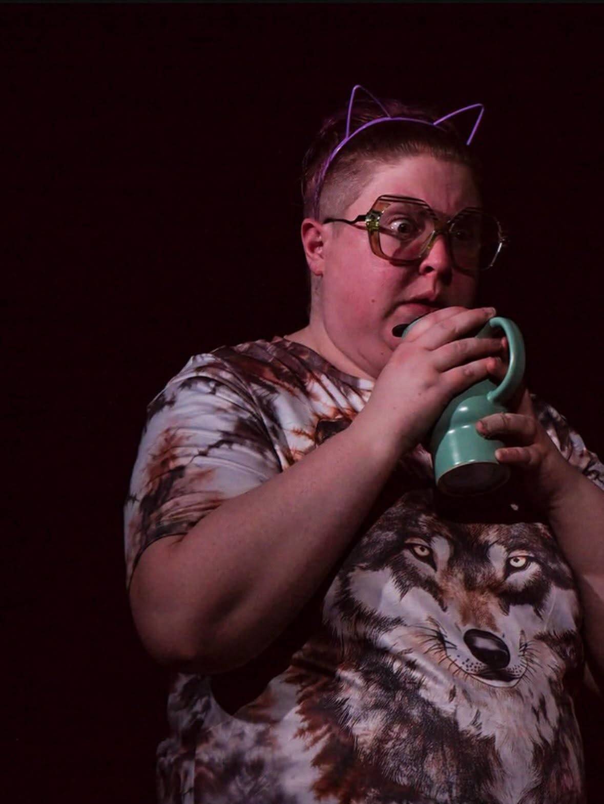 A performer in a wolf-print tie-dye shirt and cat-ear headband drinks from a mint green mug, eyes wide.