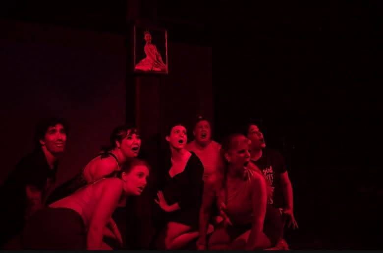 The Dance Nation company crowded together under deep red light, mouths open mid-shout, beneath a framed portrait.