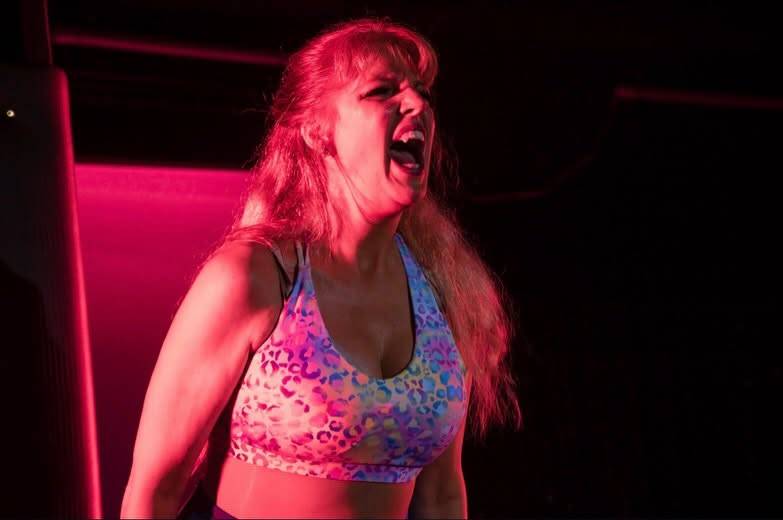 A dancer in a bright patterned sports bra throws her head back and roars under pink and red light.