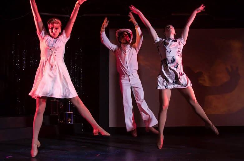 Three dancers in pale costumes caught mid-leap with their arms flung overhead.