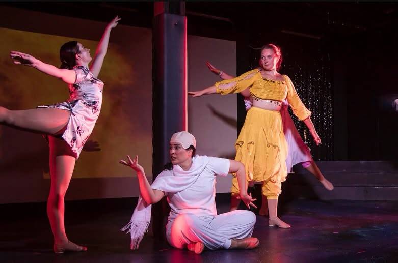 Three dancers in a held pose: one in arabesque, one seated in a white cap, one in a yellow two-piece costume.
