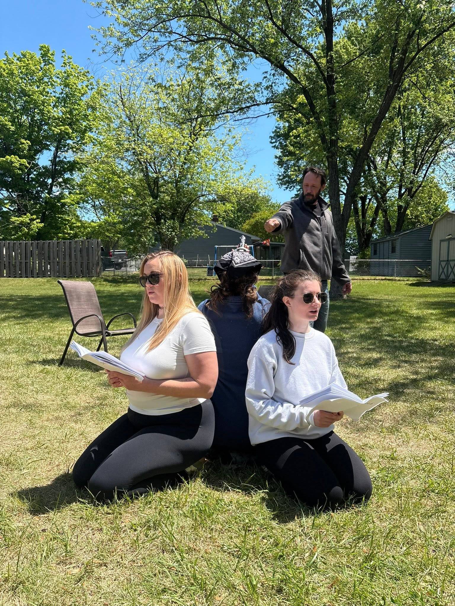 Three performers rehearse on a sunlit lawn, two kneeling back to back holding scripts and speaking, a third kneeling between them in a patterned crown. A fourth person stands behind, pointing.