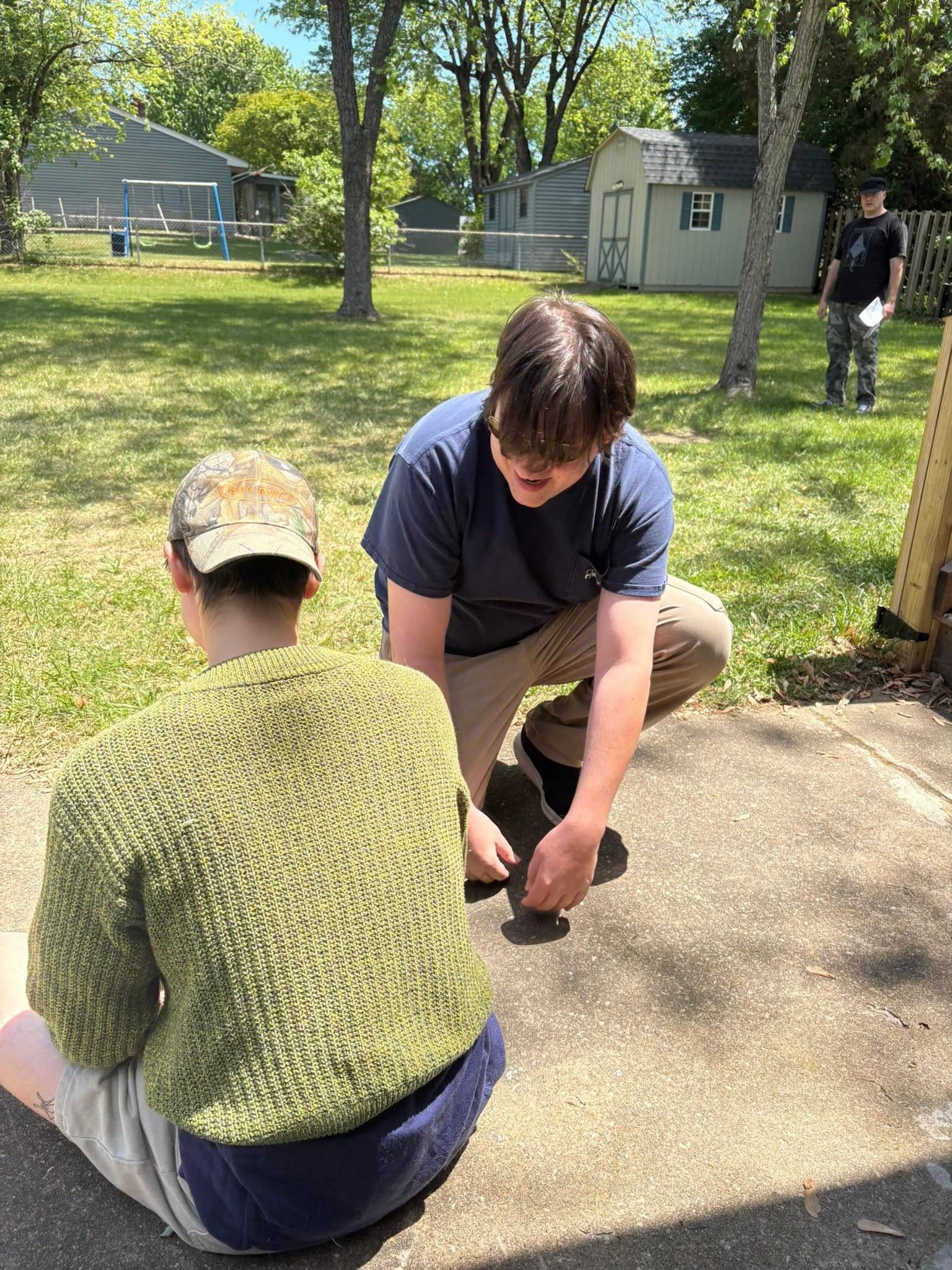 Two people crouch over a mark on a concrete path in a back garden, one reaching down to it. A third stands at the fence in the background holding a sheet of paper.