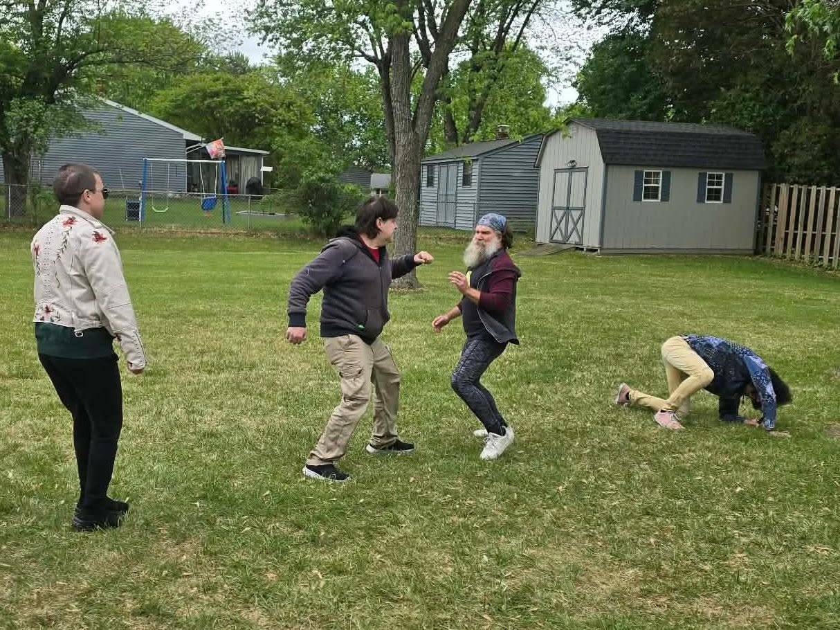 Five people rehearse a fight on a lawn: one throws a punch, another recoils, a third doubles over at the right, and two more watch from the edges.