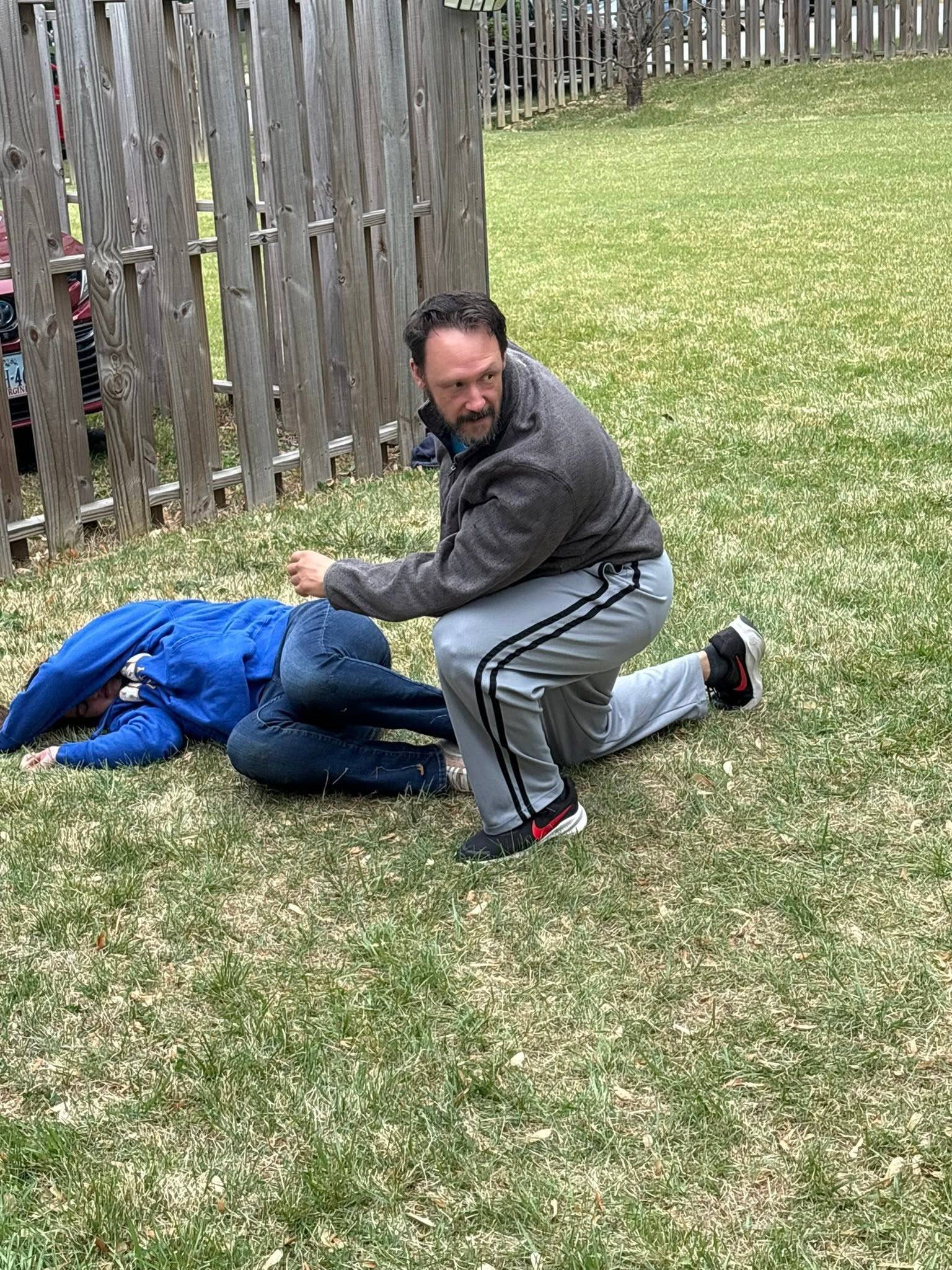 Two people rehearse a fall on the grass beside a wooden fence, one lying face down while the other kneels over them with a fist drawn back, looking toward the camera.