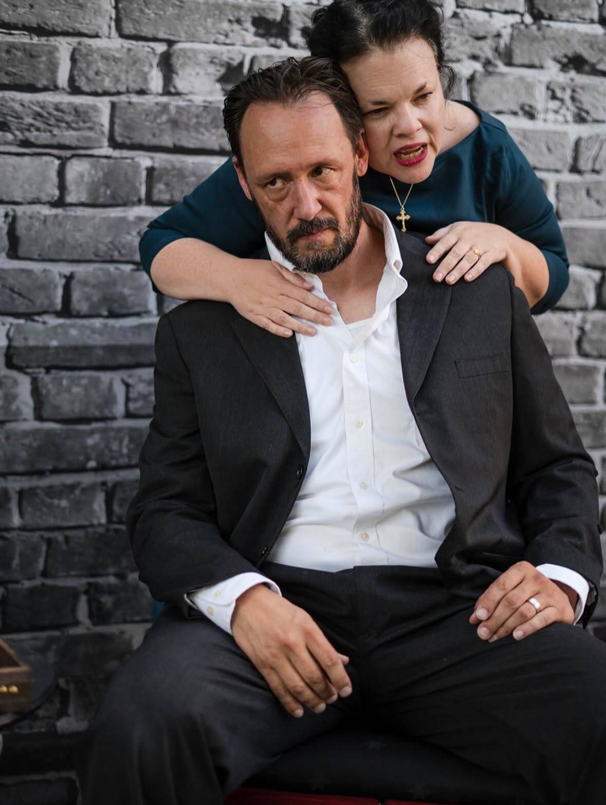 Two performers against a grey brick wall: one sits in a dark suit and open white shirt looking down; the other stands behind in a teal blouse and a gold cross, arms draped over their shoulders, speaking close to their ear.