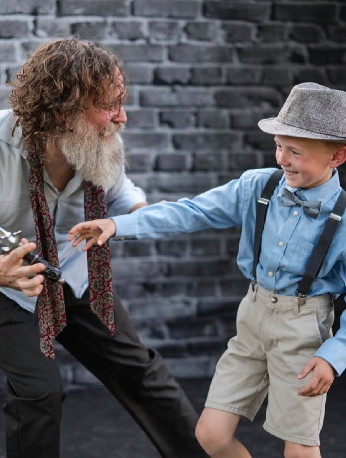 A performer with long curly hair and a full beard, in a pale shirt and a loose paisley tie, crouches holding a dagger, facing a young performer in a blue shirt, grey trilby, bow tie and braces who reaches toward them. A grey brick wall is behind.