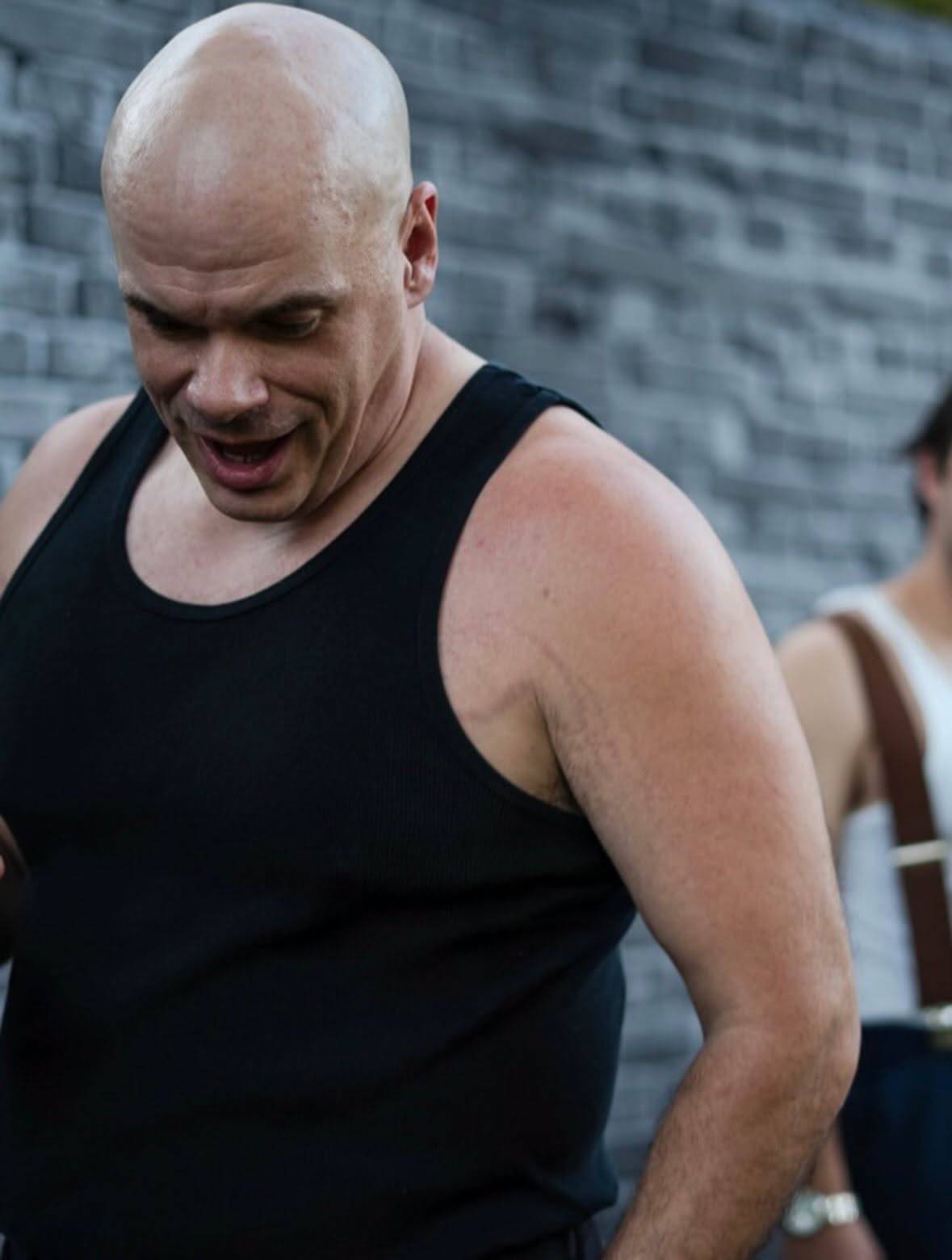 A performer in a black vest looks down and away, mouth open mid-line, against a grey brick wall. Another performer in a white vest and braces stands blurred at the right edge.