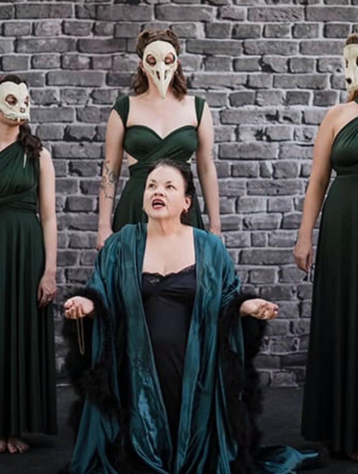 Three performers in long dark green gowns and bone-white animal-skull masks stand against a grey brick wall. In front of them a fourth performer kneels in a black slip and a teal robe trimmed with black feathers, hands open and turned upward, looking up.