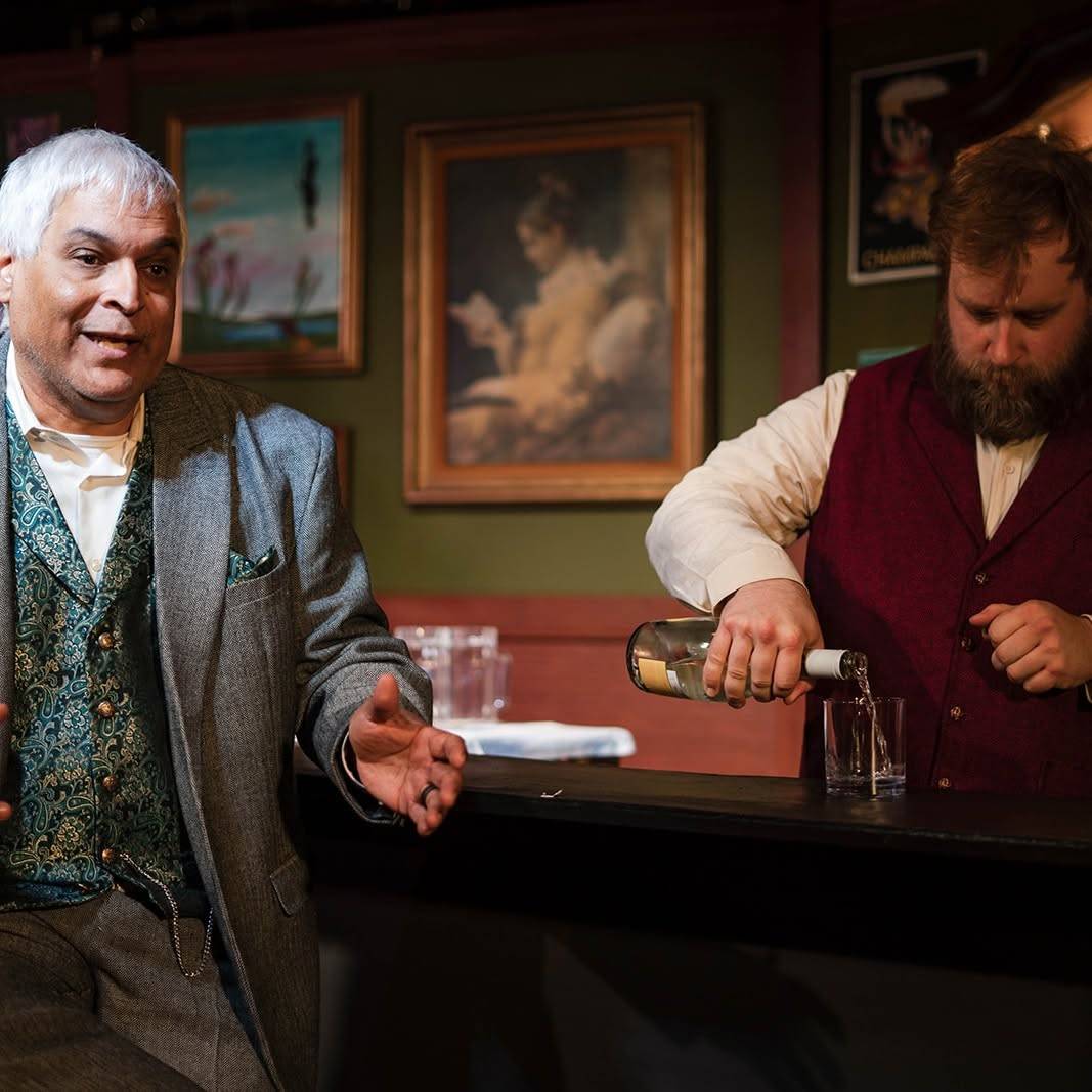 The performer stands at the bar with one hand open, speaking, while another performer in a shirtsleeves and a burgundy waistcoat pours a drink into a glass on the counter. Framed paintings hang on the green wall behind.