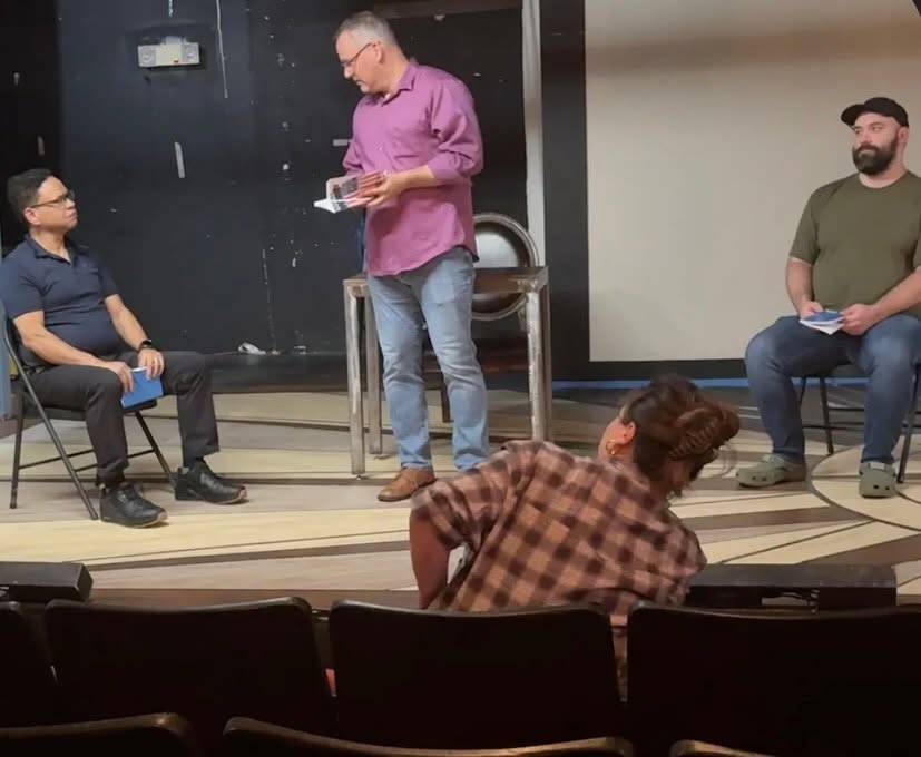 Performers sit around a rehearsal space while one person stands and reads from a script.