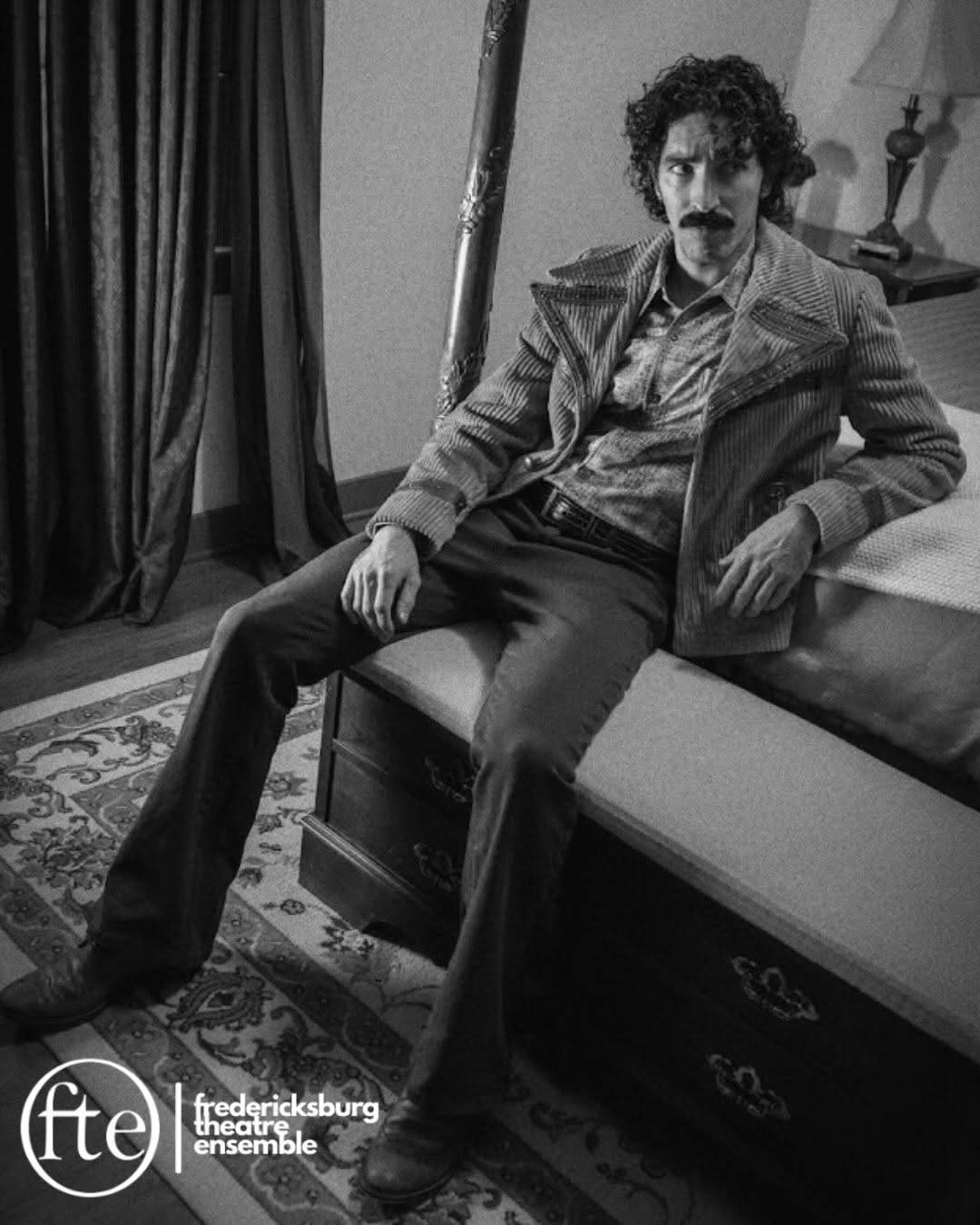A performer with dark curly hair and a mustache, in a corduroy jacket and flared trousers, sits on a chest at the foot of a four-poster bed, leaning on one arm and looking toward the camera. Black and white photo with the Fredericksburg Theatre Ensemble logo in the lower left.