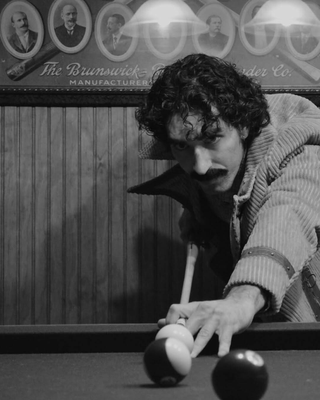 The same performer leans over a pool table lining up a shot with a cue, an eight ball in the foreground. Black and white photo.