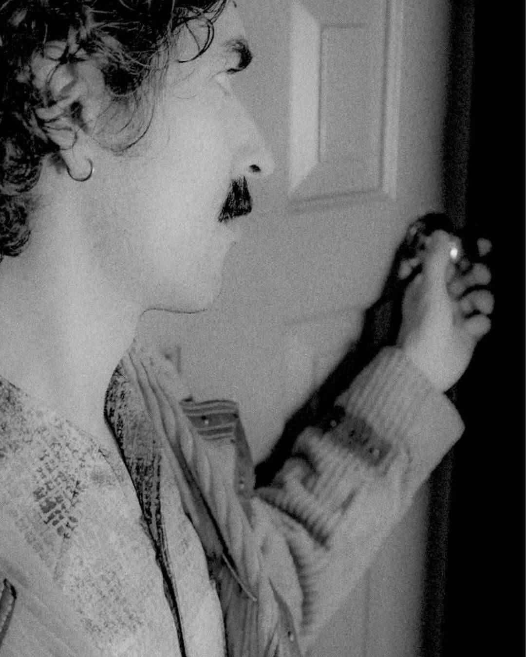 Close side view of the same performer in profile, reaching toward a door handle. Black and white photo.
