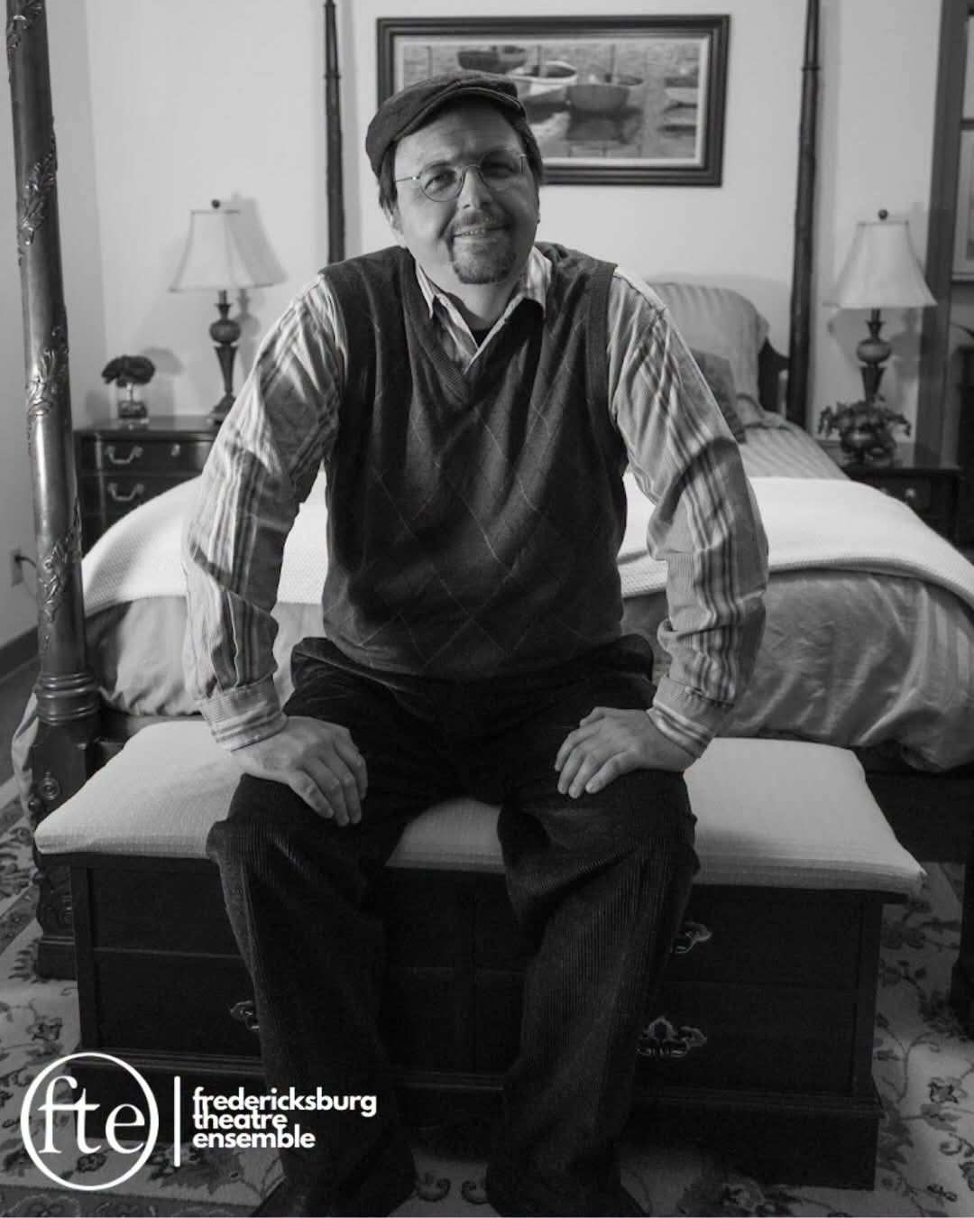 A performer in a flat cap, round glasses, argyle sweater vest and striped shirt sits on a cushioned chest at the foot of a four-poster bed, hands on their knees, with a slight smile. Black and white photo with the Fredericksburg Theatre Ensemble logo in the lower left.