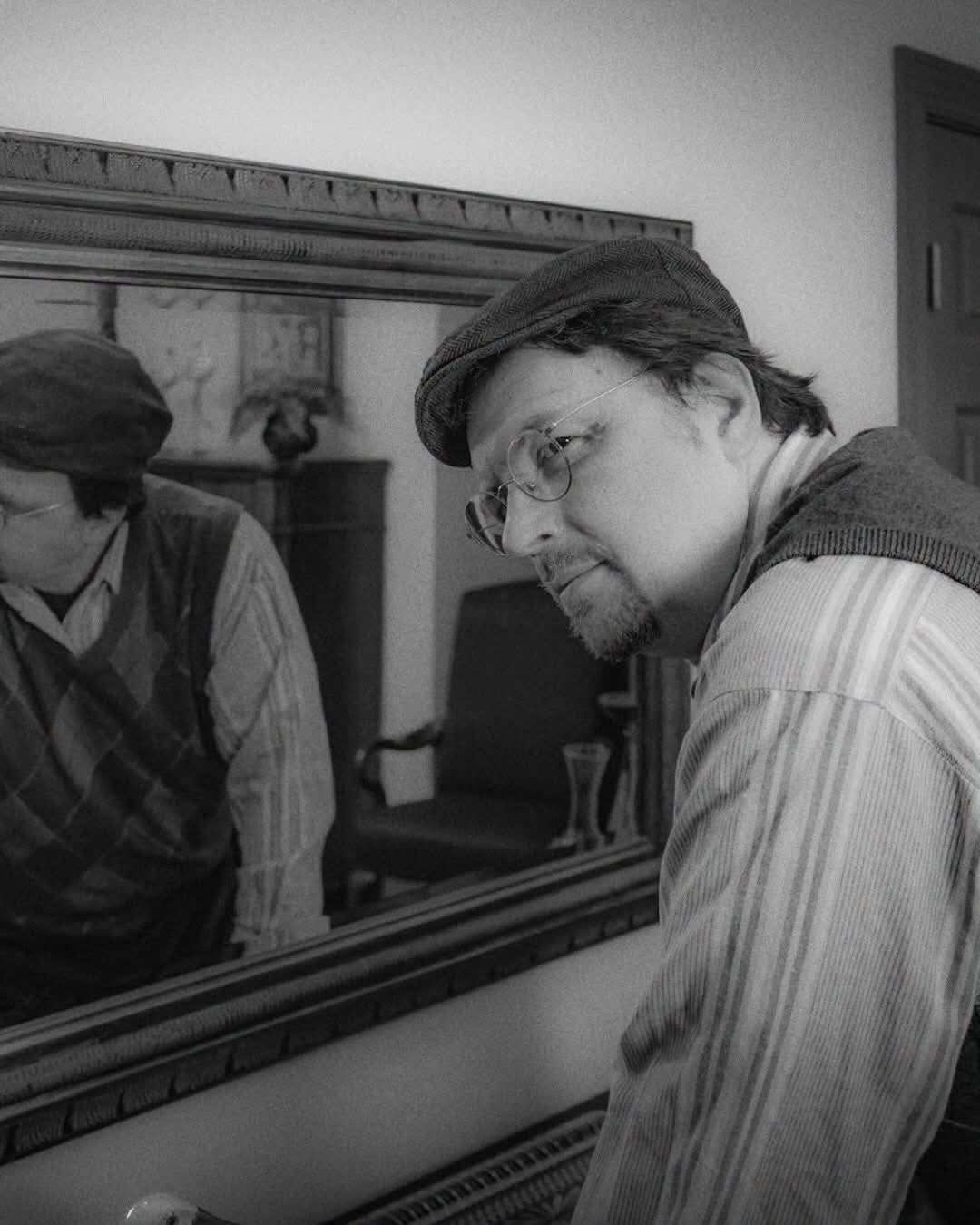 The same performer leans beside a large framed mirror that holds their reflection, looking off to the side. Black and white photo.