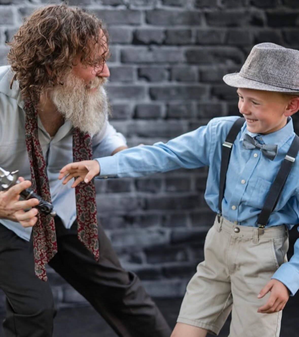 On a stage in front of a grey brick wall, a performer with long curly hair and a long white beard, in a pale shirt with a paisley tie hanging loose, crouches holding a handful of small metal objects while a child in a flat cap, bow tie, blue shirt, braces and shorts reaches toward them, laughing.