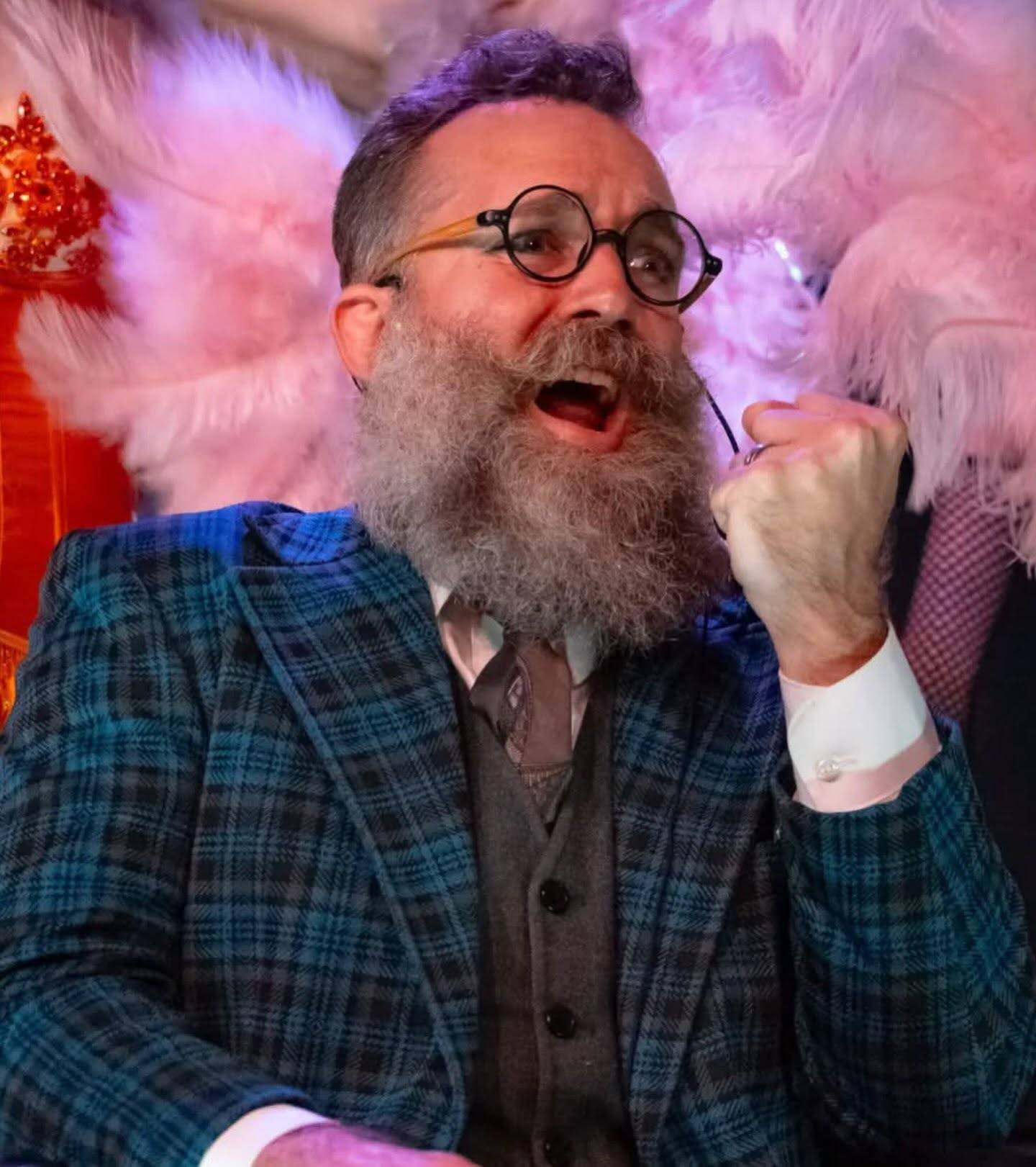 A performer with purple-lit hair, round black glasses and a long grey beard, in a blue plaid three-piece suit, mouth open mid-shout with one fist raised, framed by pink feather fans.