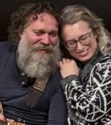 Two people sitting close together, one with a long grey beard and a jewel on their forehead holding a guitar, the other leaning their head on that shoulder with eyes closed, both smiling.