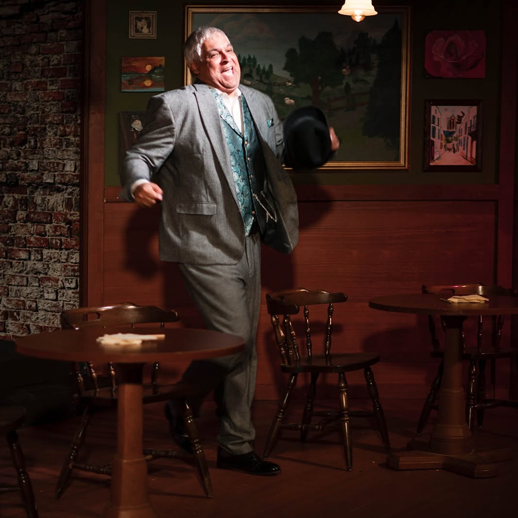 Gaston, in a grey suit and teal brocade vest, dances gleefully among the café tables of the Lapin Agile, black hat in hand.