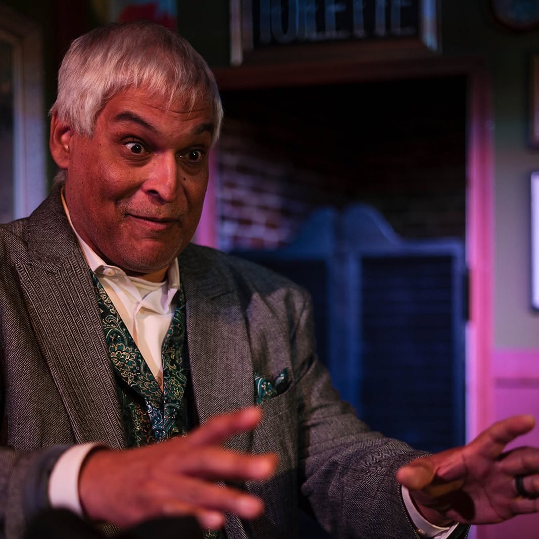 Close-up of Gaston mid-story, eyes wide and hands spread, in his herringbone suit and paisley vest.