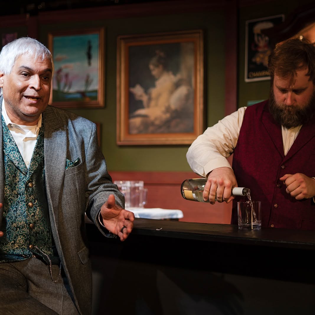 Gaston gestures from his seat at the bar while the bartender pours a drink beside him.