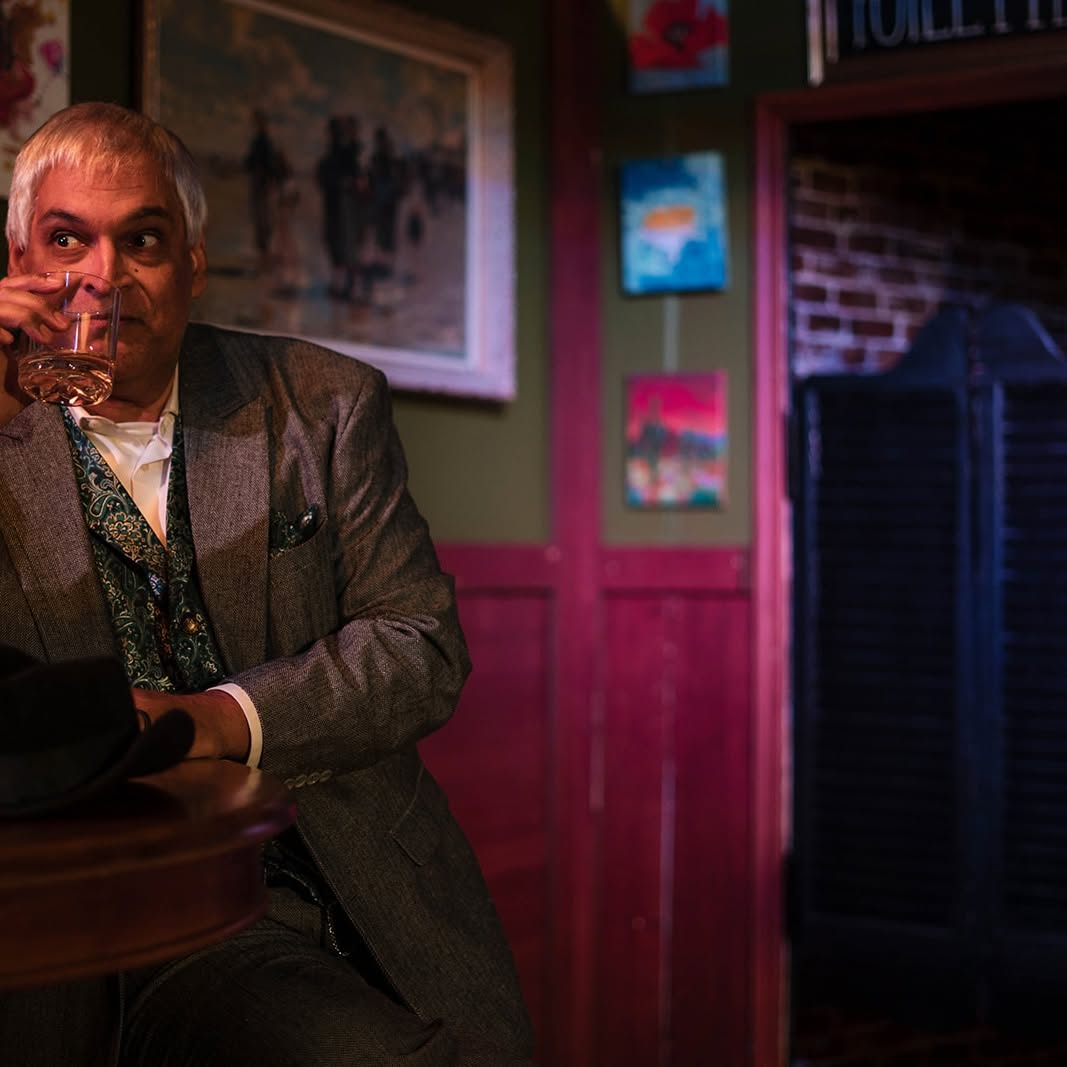Gaston pauses for a sip at a café table, hat resting beside him, glancing across the bar.