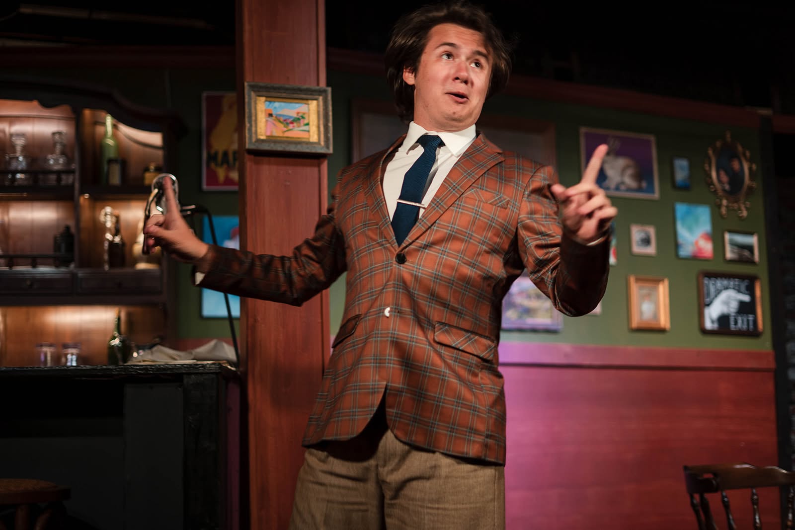Schmendiman, in a plaid suit and navy tie, holds forth with both hands raised on the Lapin Agile bar set.
